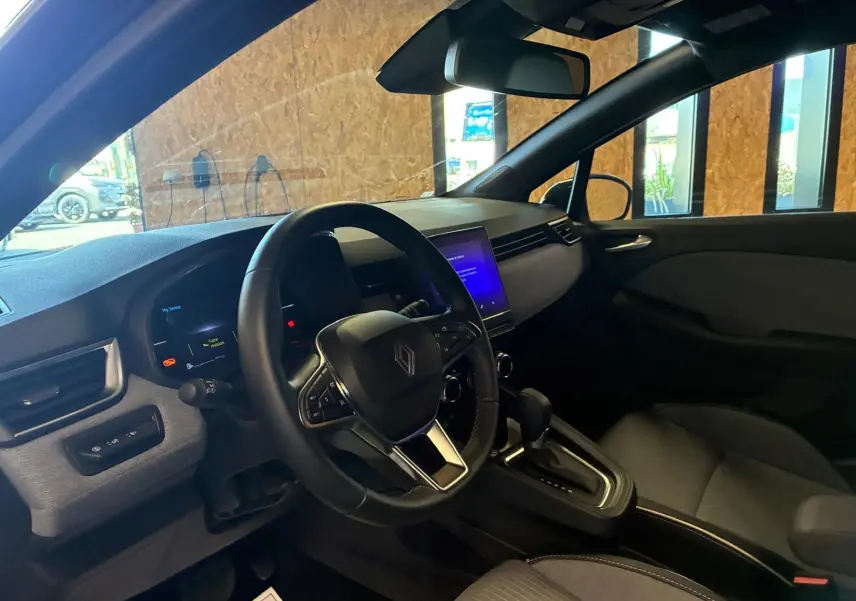 Vue intérieure côté conducteur de la Renault Clio 2025 gris schiste, avec volant multifonction et écran tactile central allumé.
