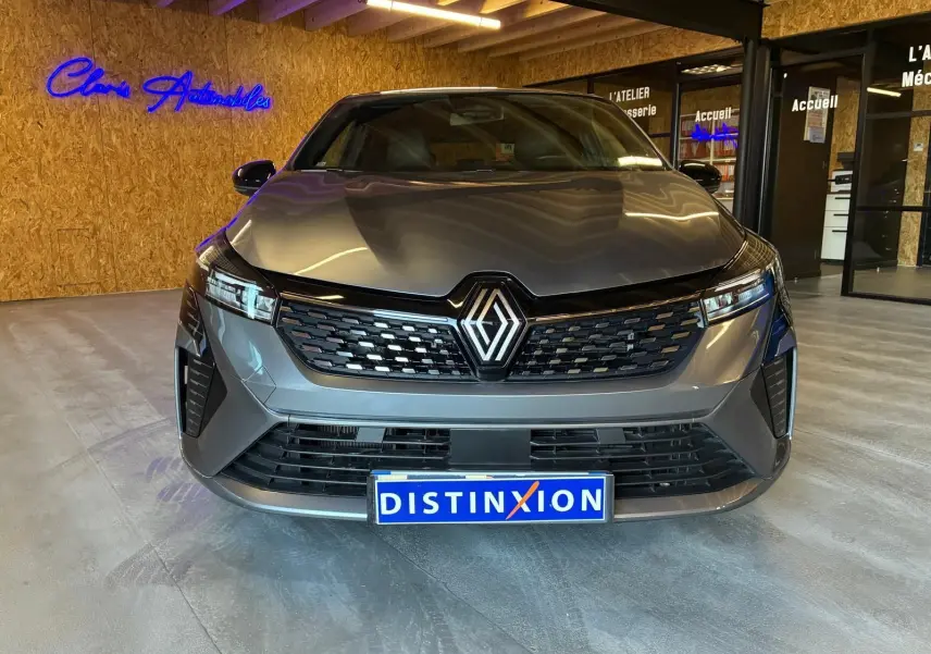 Vue frontale d'une Renault Clio 2025 gris schiste, avec calandre noire et logo Renault lumineux, en intérieur showroom.