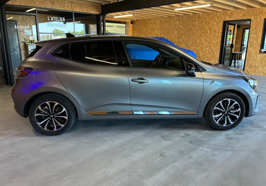 Profil côté gauche de la Renault Clio 2025 gris schiste, avec jantes noires et détails chromés, en intérieur d'atelier.