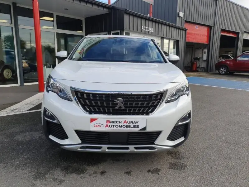 Vue de face d'un Peugeot 3008 blanc nacré avec calandre noire et logo lion au centre, stationné devant un garage.