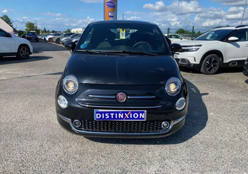 Vue frontale d'une Fiat 500 noire 2024 avec phares allumés et plaque Distinxion sur un parking extérieur.