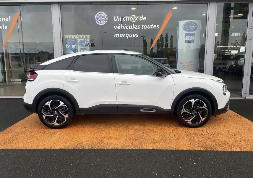 Profil droit d'une Citroën C4 blanche 2022 avec toit noir et jantes alliage distinctives, stationnée devant une vitrine.