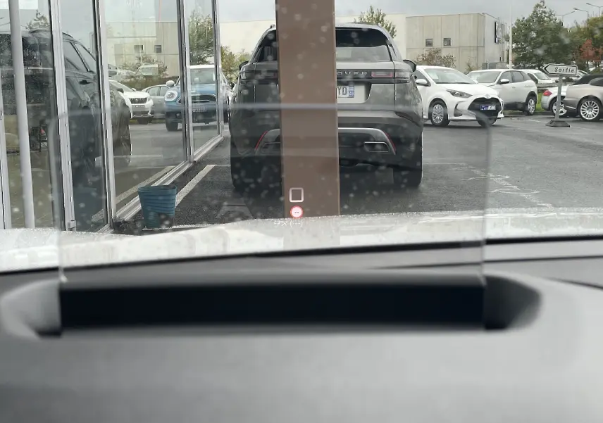 Vue depuis l'intérieur d'un véhicule blanc, montrant le pare-brise avec affichage tête haute et un SUV noir garé devant.