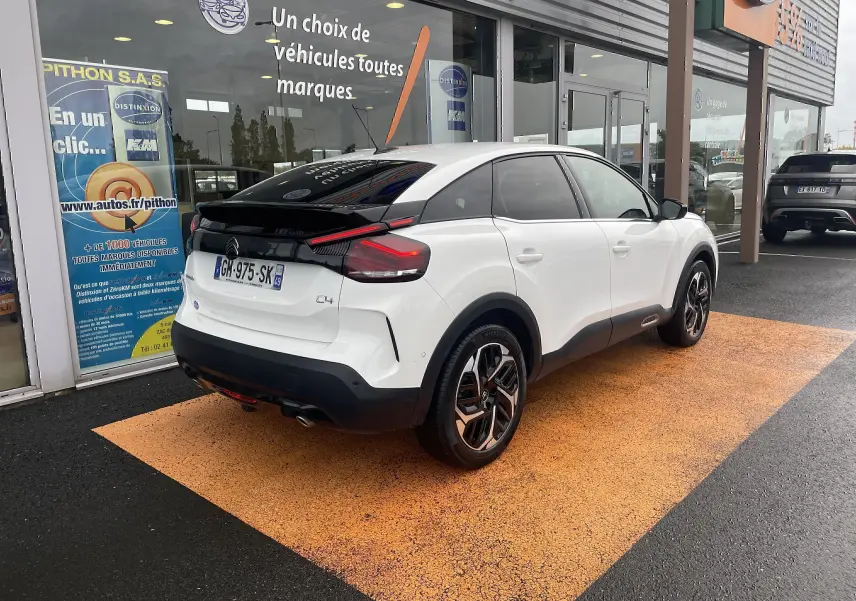 Vue 3/4 arrière droite d'une Citroën C4 blanche 2022 avec toit noir, feux LED et jantes bicolores, stationnée devant un showroom.