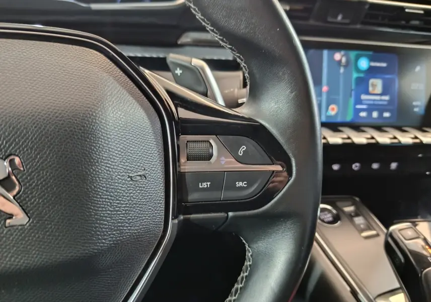 Gros plan sur le volant cuir noir de la Peugeot 508 gris clair, avec commandes audio et écran tactile en arrière-plan.