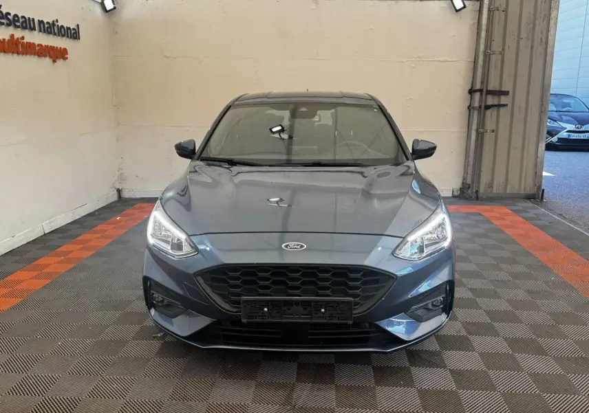 Vue frontale d'une Ford Focus 5 portes bleu métallisé avec calandre noire et phares allumés.