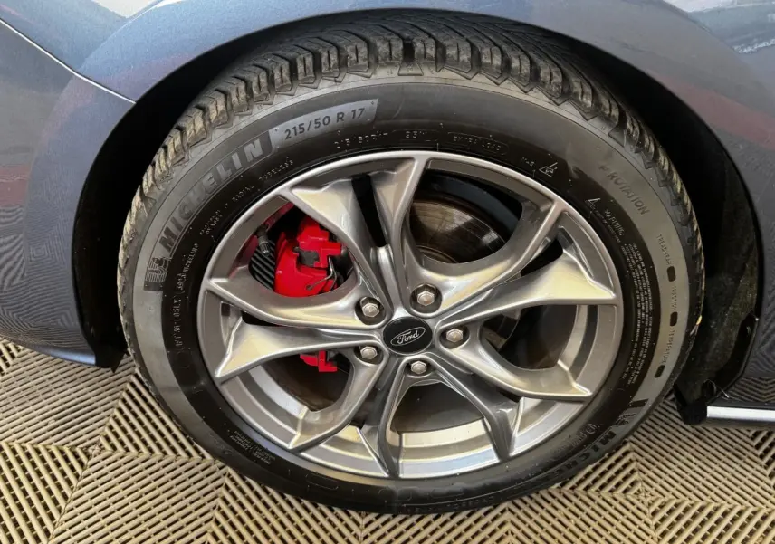 Gros plan sur la roue avant droite d’une Ford Focus bleu, jante alliage grise avec étrier de frein rouge visible.