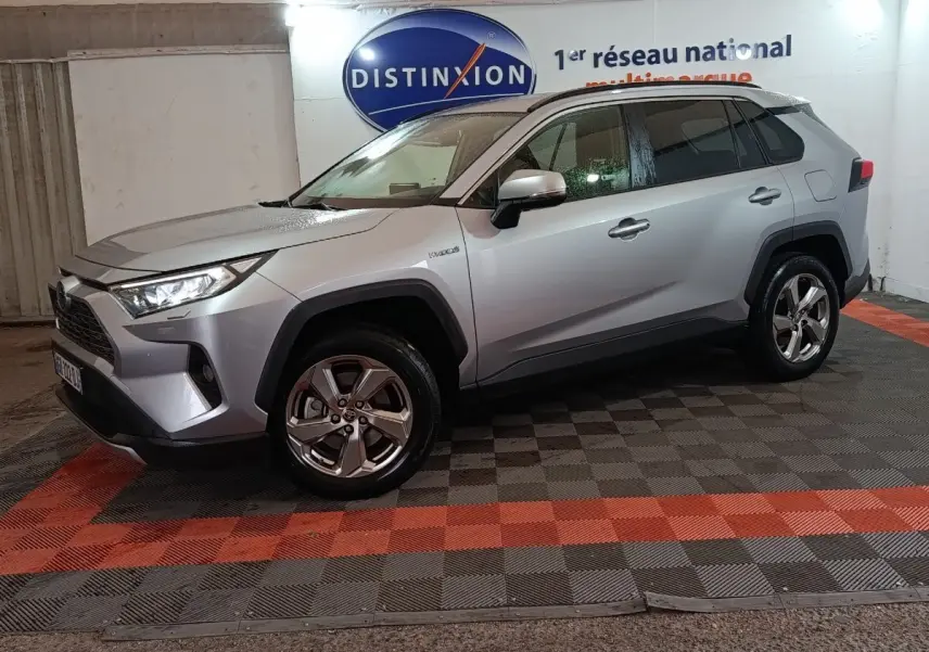 Toyota RAV4 Hybride gris clair vu de profil droit en intérieur, avec jantes alliage et logo Distinxion en arrière-plan.