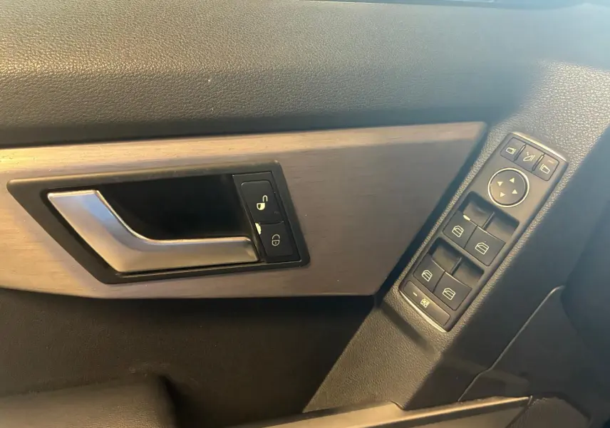 Gros plan sur la poignée de porte côté gauche et les commandes de vitres électriques d’un Mercedes GLK noir, intérieur bois et cuir.