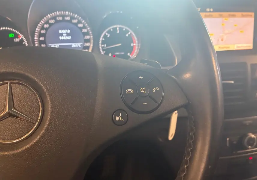Gros plan sur le volant en cuir noir d’un Mercedes GLK 220 CDI 2012, avec commandes audio et écran GPS en arrière-plan.