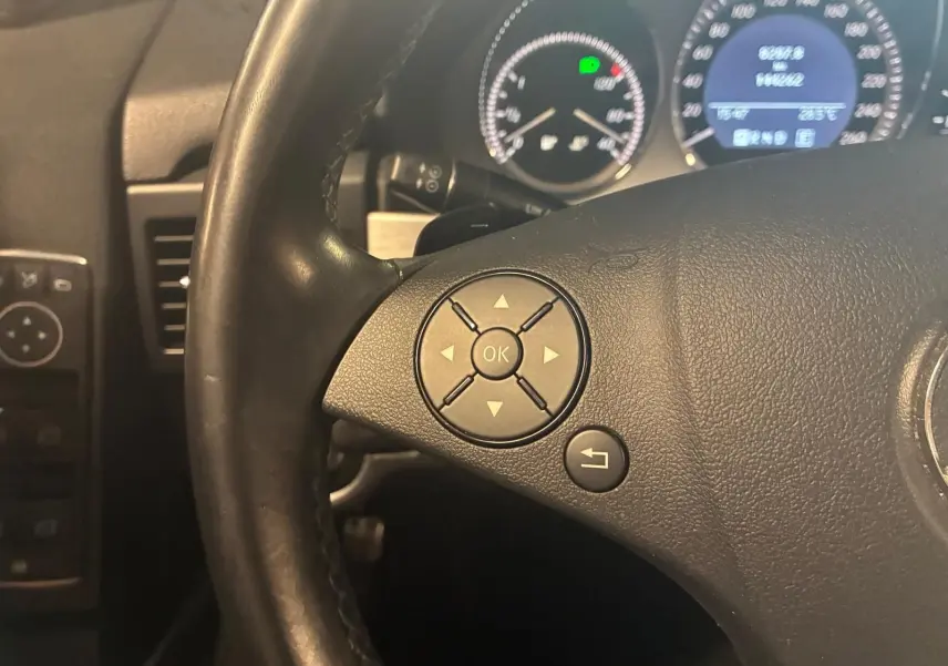 Gros plan sur le volant en cuir noir d’un Mercedes GLK 220 CDI, avec commandes intégrées et tableau de bord éclairé en arrière-plan.