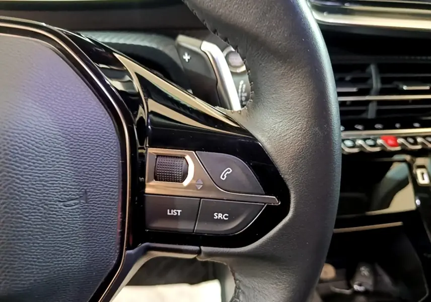 Gros plan sur le volant cuir noir de la Peugeot 2008, avec commandes multifonctions et tableau de bord en arrière-plan.