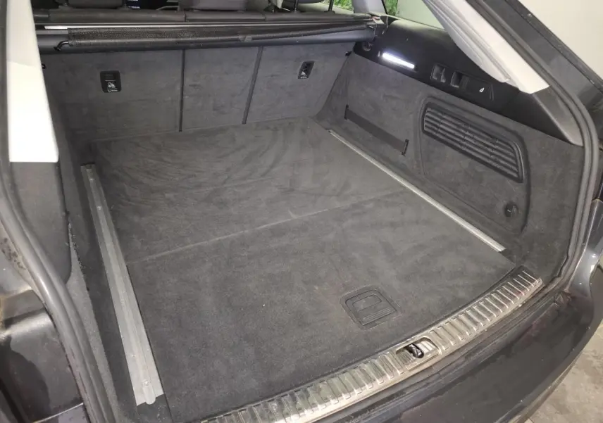 Coffre ouvert vu de l'arrière d'une Audi A6 Avant gris, avec tapis de sol noir et finitions métalliques visibles.