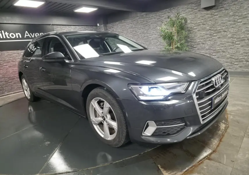 Audi A6 Avant gris métallisé vue 3/4 avant droit avec phares LED allumés en intérieur showroom.