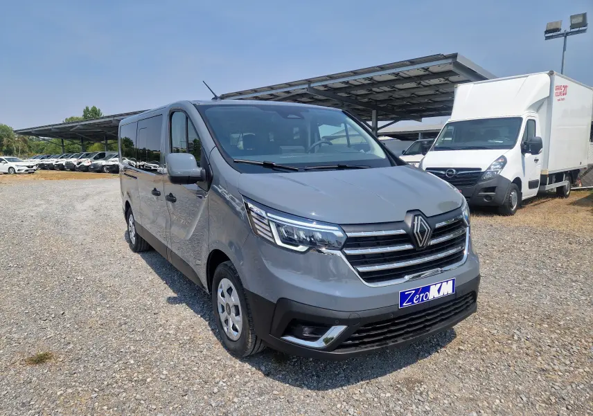 Vue 3/4 avant droit d’un Renault Trafic Combi gris urbain stationné sur un sol gravillonné avec un abri solaire en arrière-plan.