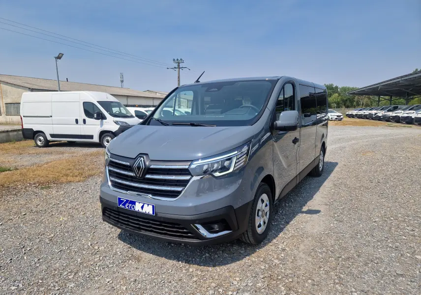 Renault Trafic Combi gris urbain vu en 3/4 avant droit sur un terrain gravillonné avec plusieurs véhicules en arrière-plan.
