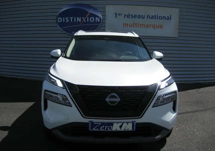 Vue de face du Nissan X-TRAIL blanc nacré avec calandre noire et phares LED allumés sur fond de façade commerciale.