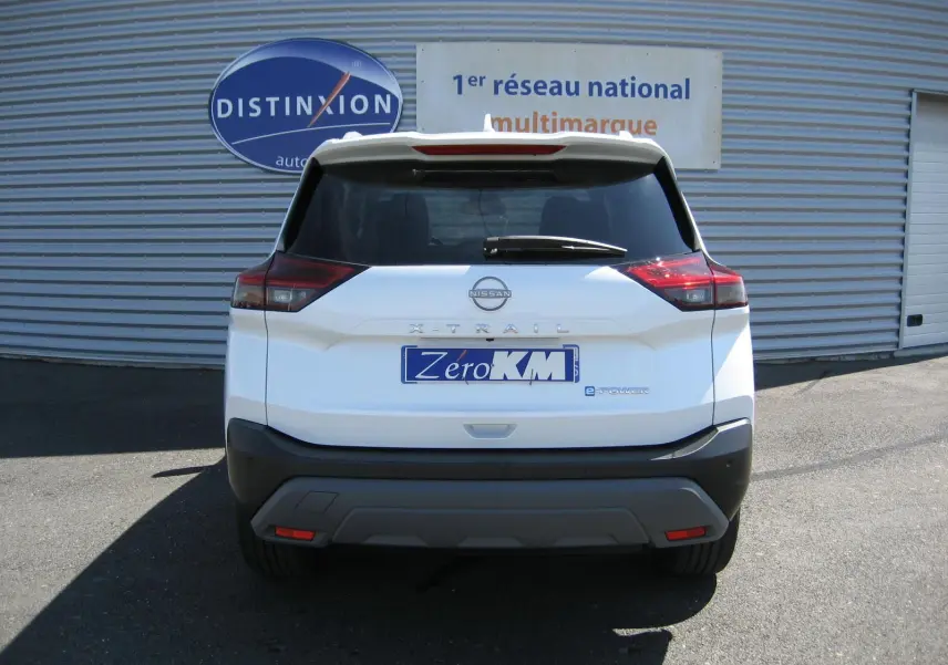 Vue arrière d'un Nissan X-Trail blanc nacré avec logo e-Power et plaque "Zéro KM" devant un mur métallique.