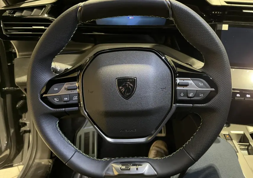 Volant cuir noir avec surpiqûres vertes et logo GT au centre d'une Peugeot 308 Gris Selenium, vue de face intérieure.