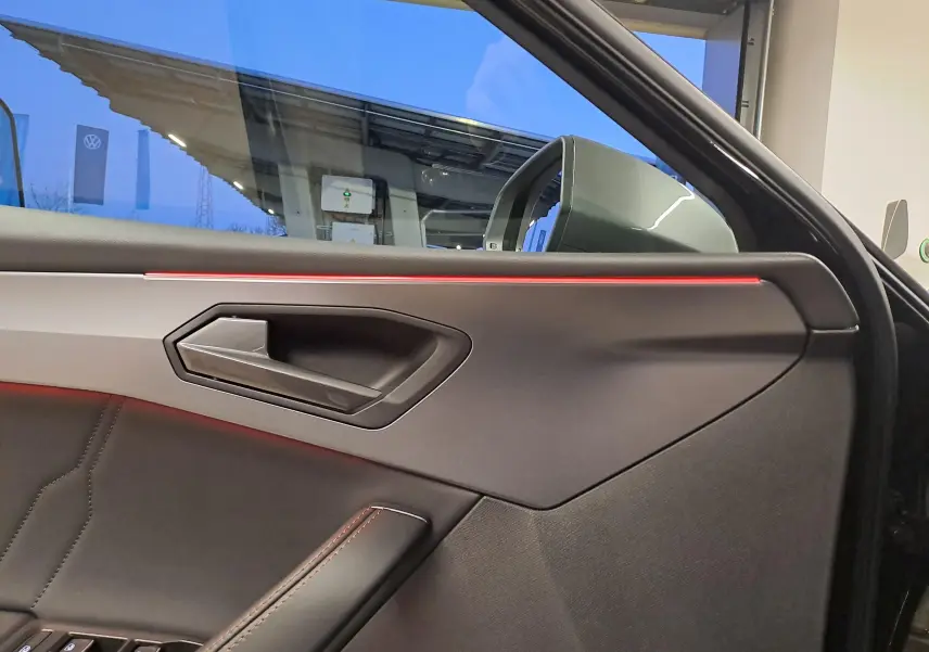 Vue intérieure côté gauche de la porte d’une CUPRA Leon 2023 noir minuit avec éclairage d’ambiance rouge et poignée moderne.