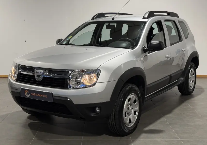Dacia Duster gris en 3/4 avant droit avec attelage, phares allumés et protections noires autour des roues.