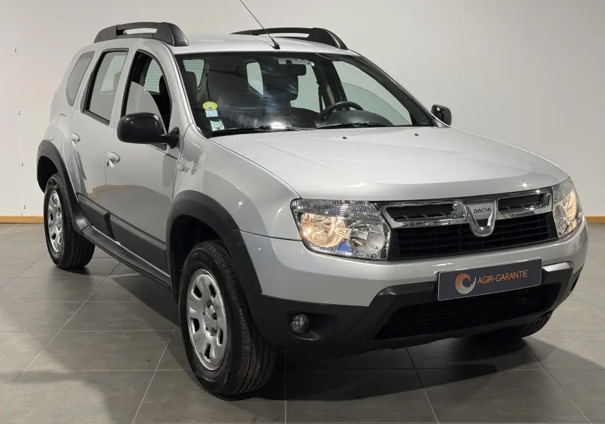Vue 3/4 avant droit d'un Dacia Duster gris 2011 avec attelage et protections noires sur les passages de roues.