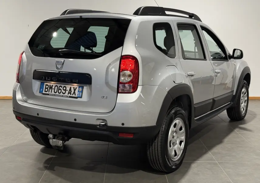Vue 3/4 arrière droite d'un Dacia Duster gris 2011 avec attelage et protections noires sur les passages de roues.