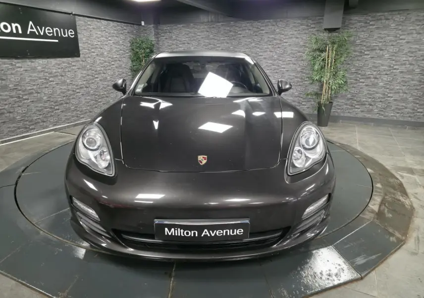 Vue avant d'une Porsche Panamera marron métallisé avec phares ovales et plaque Milton Avenue en intérieur showroom.
