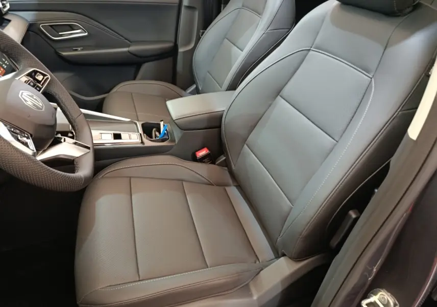 Vue intérieure côté conducteur du MG Motor ZS 1.5L Hybrid+ Luxury 2026, sièges cuir gris foncé avec accoudoir central.