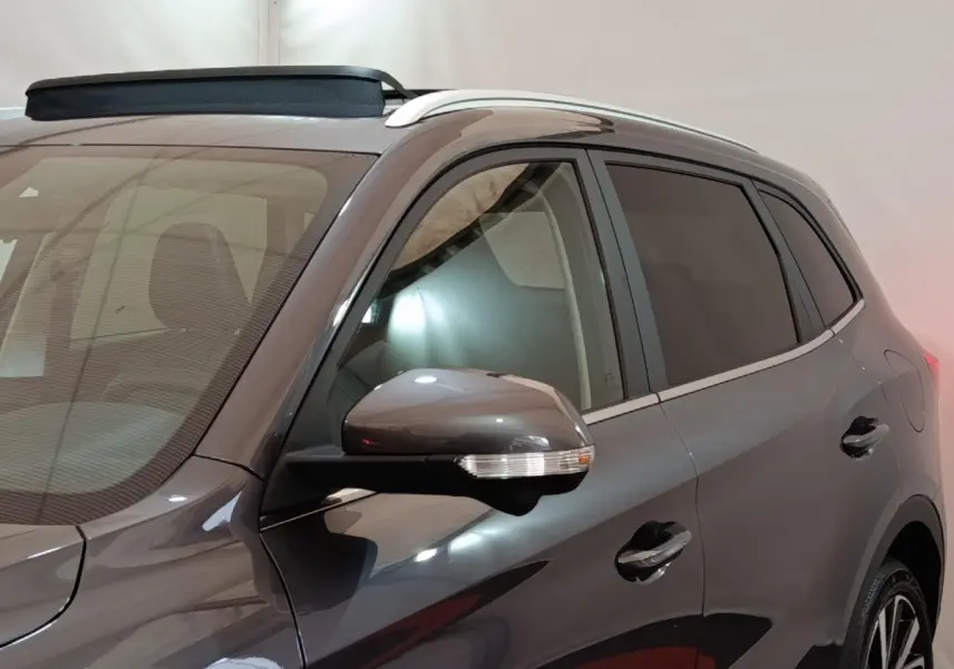 Vue de côté droit avant d’un MG ZS gris foncé avec rétroviseur et barres de toit visibles dans un intérieur neutre.