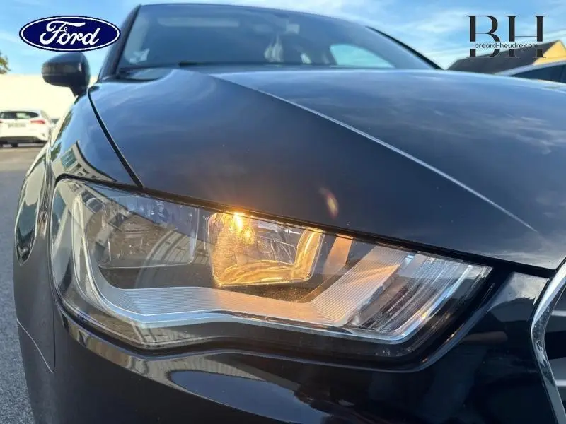 Gros plan sur le phare avant gauche d'une Audi A3 Sportback noire brillant, mettant en valeur son design moderne.