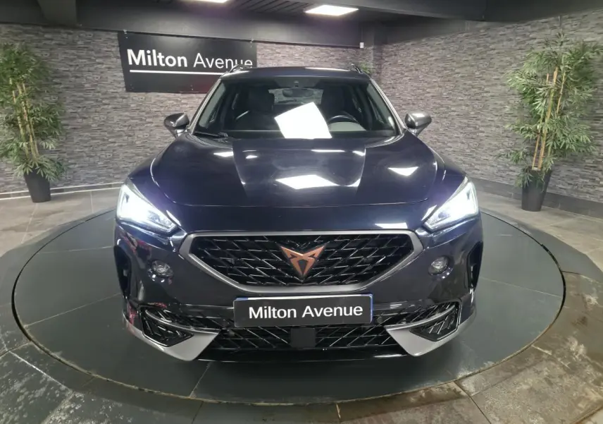 Vue frontale d'un CUPRA Formentor bleu avec calandre noire et logo cuivre, phares LED allumés dans un showroom.