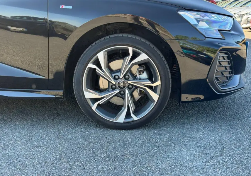 Gros plan sur la roue avant droite et le pare-chocs avant d’une Audi A3 Sportback noir Mythic métallisé avec jante alu distinctive.