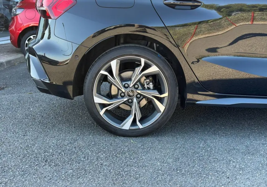 Gros plan sur la roue arrière droite d’une Audi A3 Sportback noire Mythic métallisée avec jante alu distinctive.