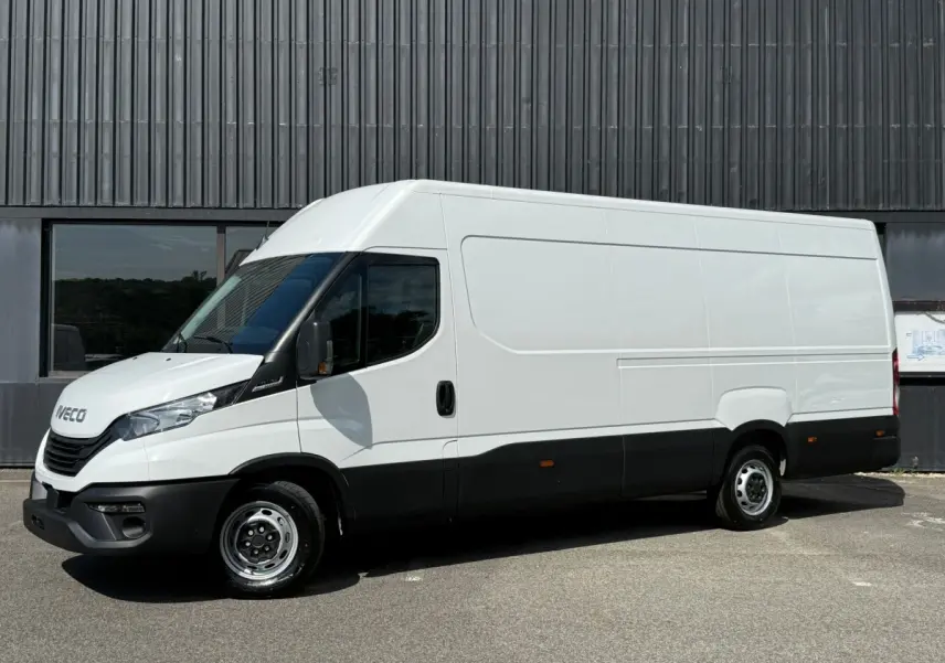 Fourgon Iveco Daily blanc vu de profil côté gauche, avec pare-chocs et bas de caisse noirs, stationné devant un bâtiment industriel.