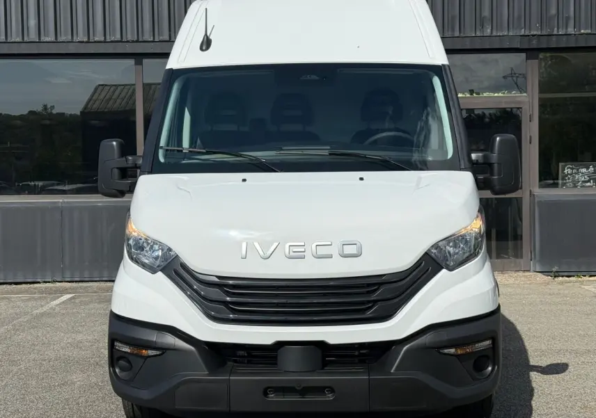 Vue frontale d’un utilitaire Iveco Daily blanc avec calandre noire et phares allumés, stationné devant un bâtiment.