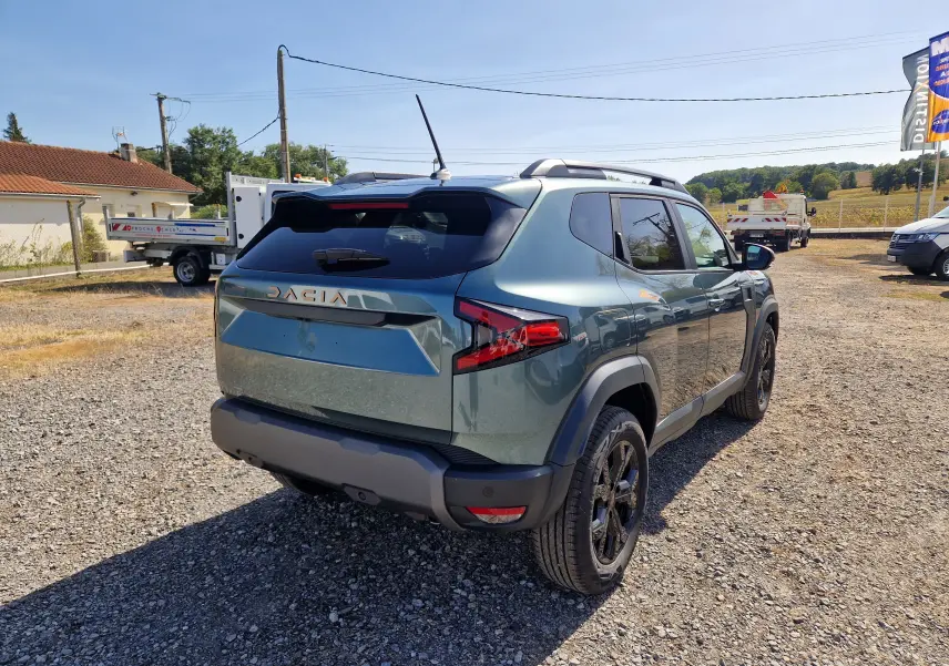 Vue 3/4 arrière droite du Dacia Duster 1.8 Hybrid 155 Vert Cèdre avec jantes noires et barres de toit visibles.