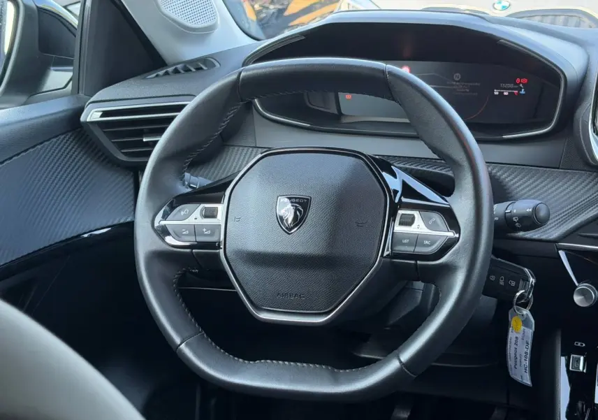 Vue rapprochée du volant compact noir avec logo Peugeot au centre et tableau de bord numérique en arrière-plan.