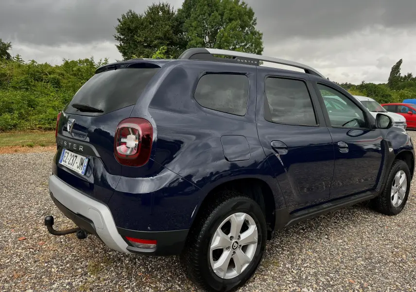Vue 3/4 arrière droite d'un Dacia Duster bleu marine 2018 avec barres de toit et attelage visible sur un sol gravillonné.