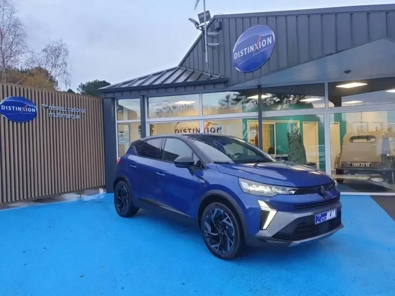 Renault Captur 2025 Bleu Iron en 3/4 avant droit, avec jantes alu noires et feux LED allumés, devant un showroom Distinxion.