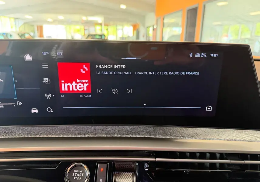 Vue rapprochée de l'écran HD 21’’ du Peugeot 5008 Hybrid 145 GT affichant France Inter, avec bouton start/stop en dessous.