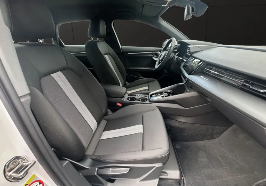 Vue intérieure côté gauche de l’Audi A3 2024 blanche, montrant les sièges tissu gris avec bande centrale claire et tableau de bord moderne.