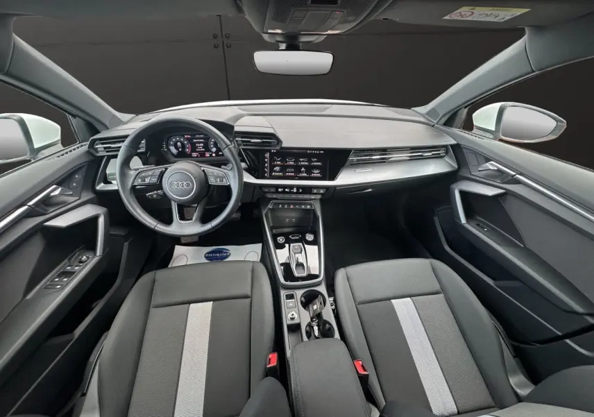 Vue intérieure avant de l'Audi A3 2024 blanche, avec tableau de bord numérique et sièges noirs à bande grise centrale.