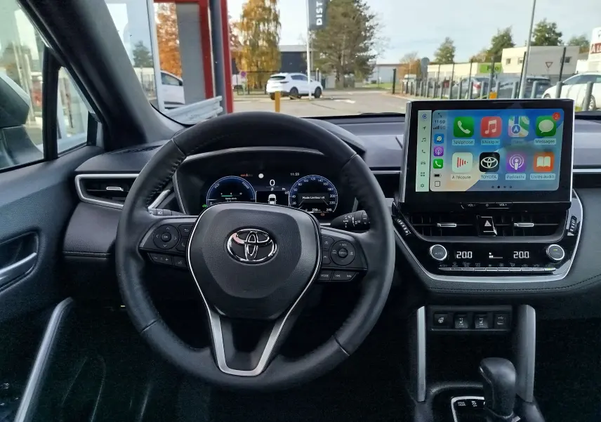 Vue intérieure du tableau de bord et volant noir d'une Toyota Corolla Cross 1.8 Hybrid 2025 avec écran tactile allumé.