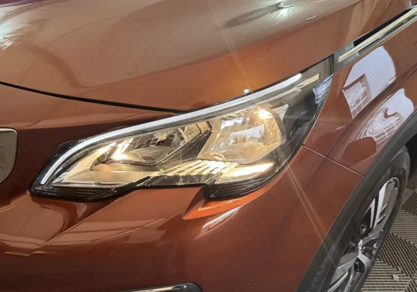 Gros plan sur le phare avant droit allumé d’un Peugeot 3008 orange métallisé, avec partie de la roue visible.