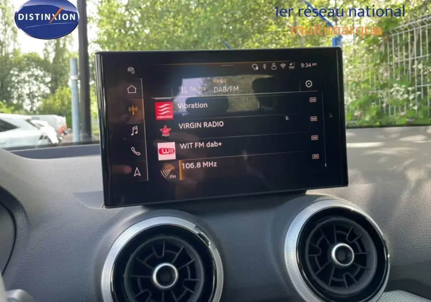Écran central tactile de l'Audi Q2 30 TDI S-Line 2025, affichant les stations radio, avec aérateurs ronds en dessous.