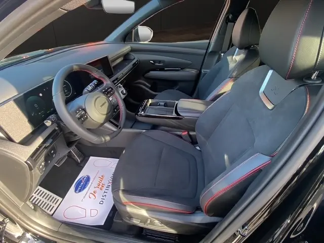 Vue intérieure côté conducteur du Hyundai Tucson 1.6 Hybrid noir, sièges en cuir et alcantara avec surpiqûres rouges et tableau de bord digital.