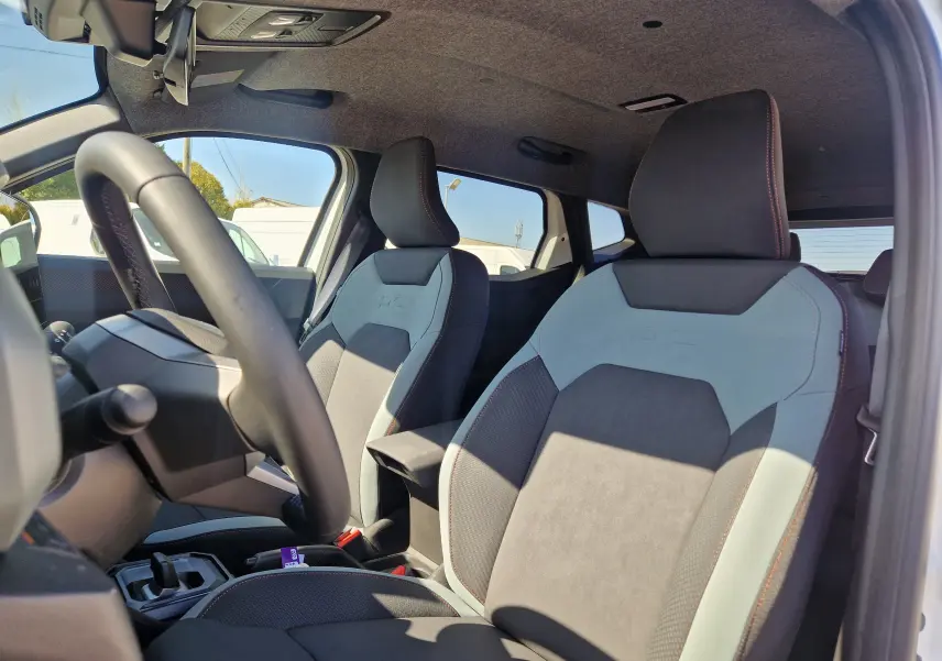 Vue intérieure côté conducteur du Dacia Duster 2026 blanc, mettant en valeur les sièges bi-matière avec surpiqûres orange.