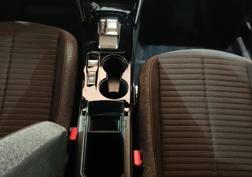 Vue intérieure centrée sur la console centrale noire brillante et les sièges tissu gris foncé avec surpiqûres rouges du Peugeot E-2008 électrique.