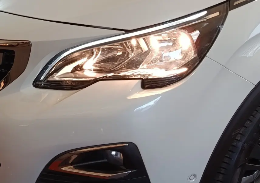Gros plan sur le phare avant droit allumé d’un Peugeot 3008 blanc, avec antibrouillard et partie de la roue visible.